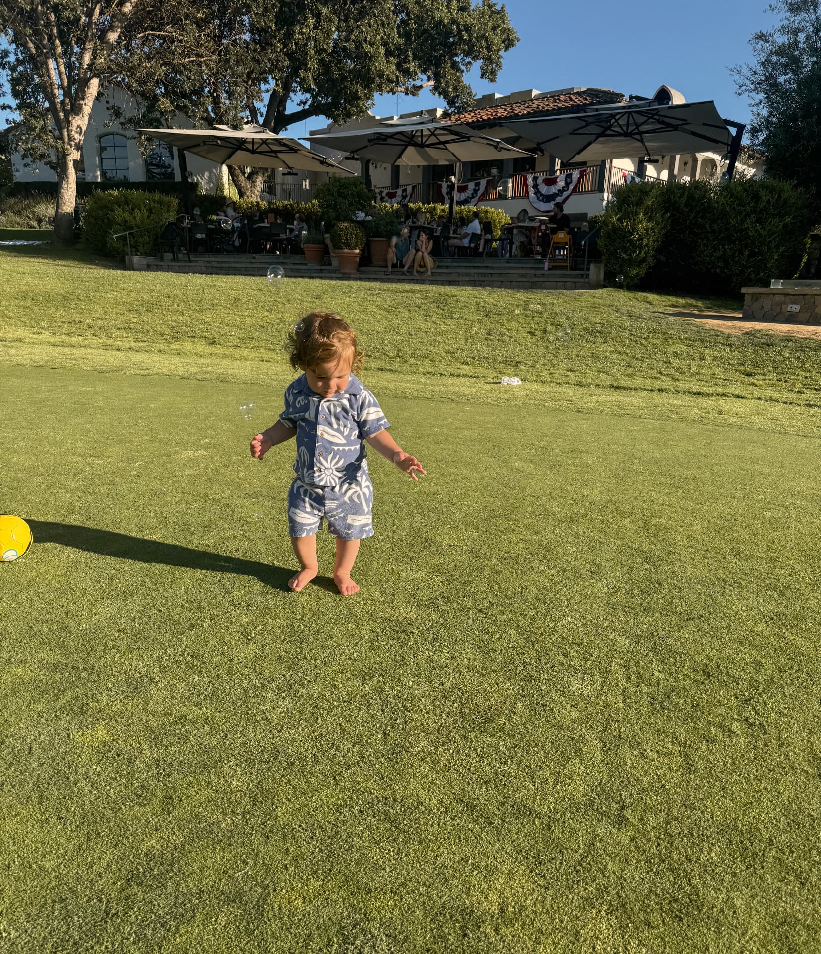 A barefoot toddler standing on the big lawn at Ojai Valley Inn with a yellow ball nearby and the terrace of Jimmy's Pub behind him
