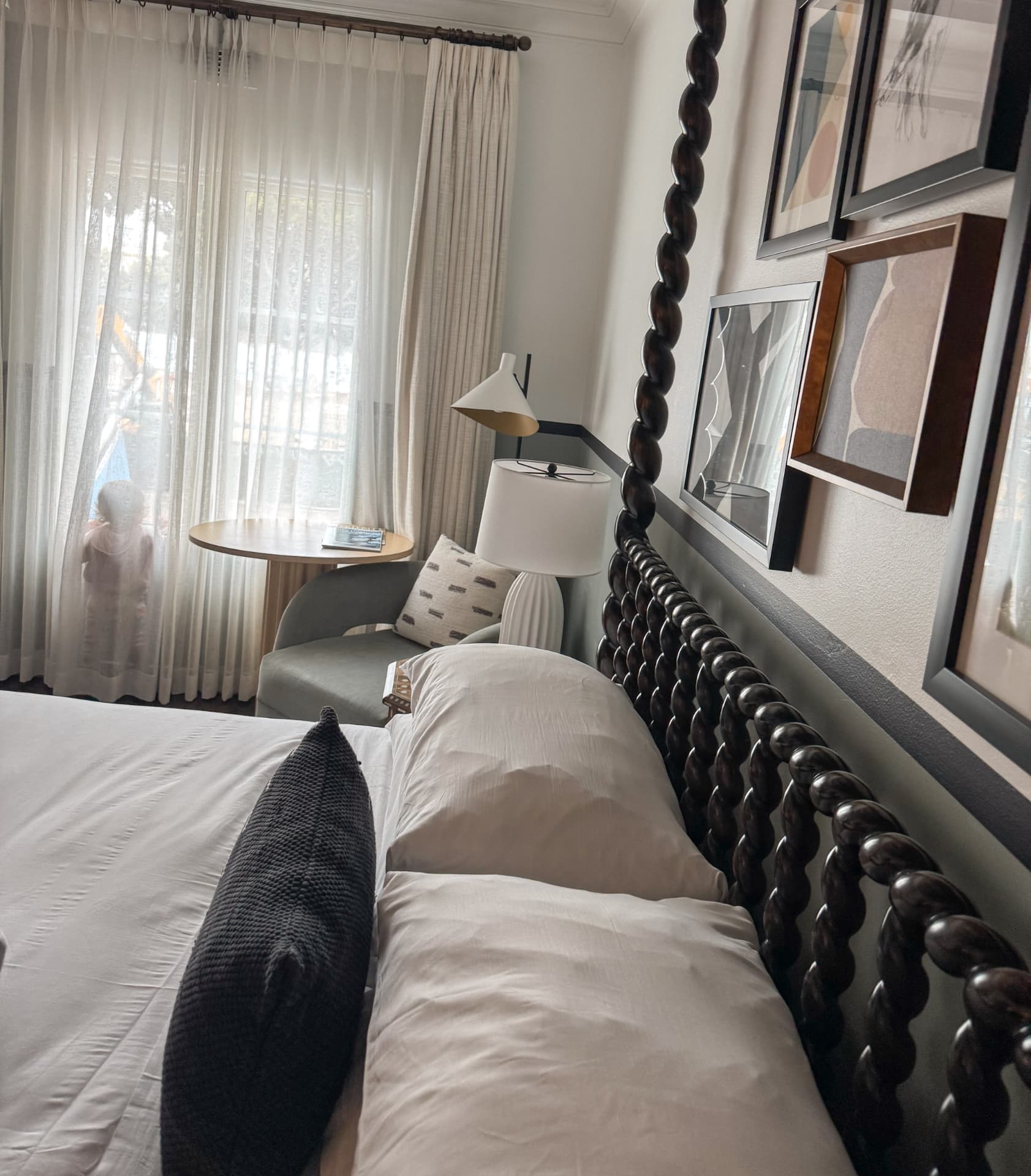 A guest room at the Kimpton Canary Hotel with a large dark spindle four-poster bed, white bedding, a green armchair and a small table by the curtained window