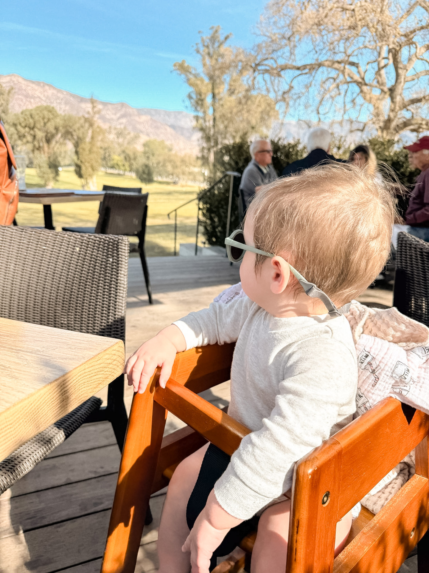 A toddler in sunglasses sitting in a high chair on an outdoor terrace at lunchtime