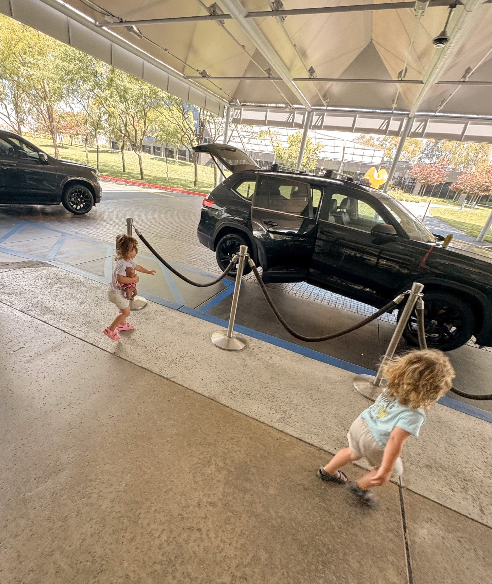 Two small children walking away across a covered hotel arrival area beside a black car with its doors open