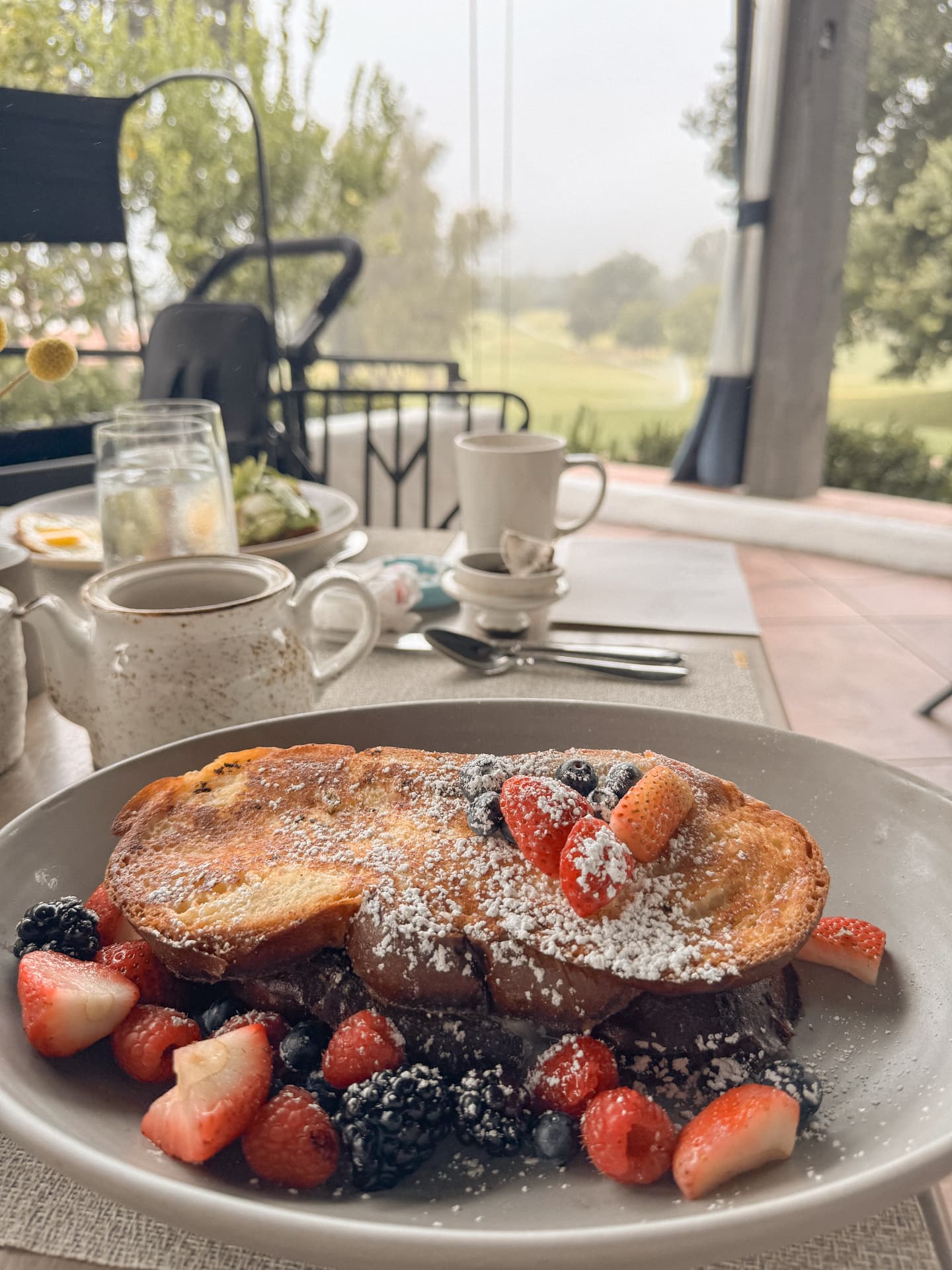 A plate of berry-topped French toast on a terrace table looking out over misty green grounds