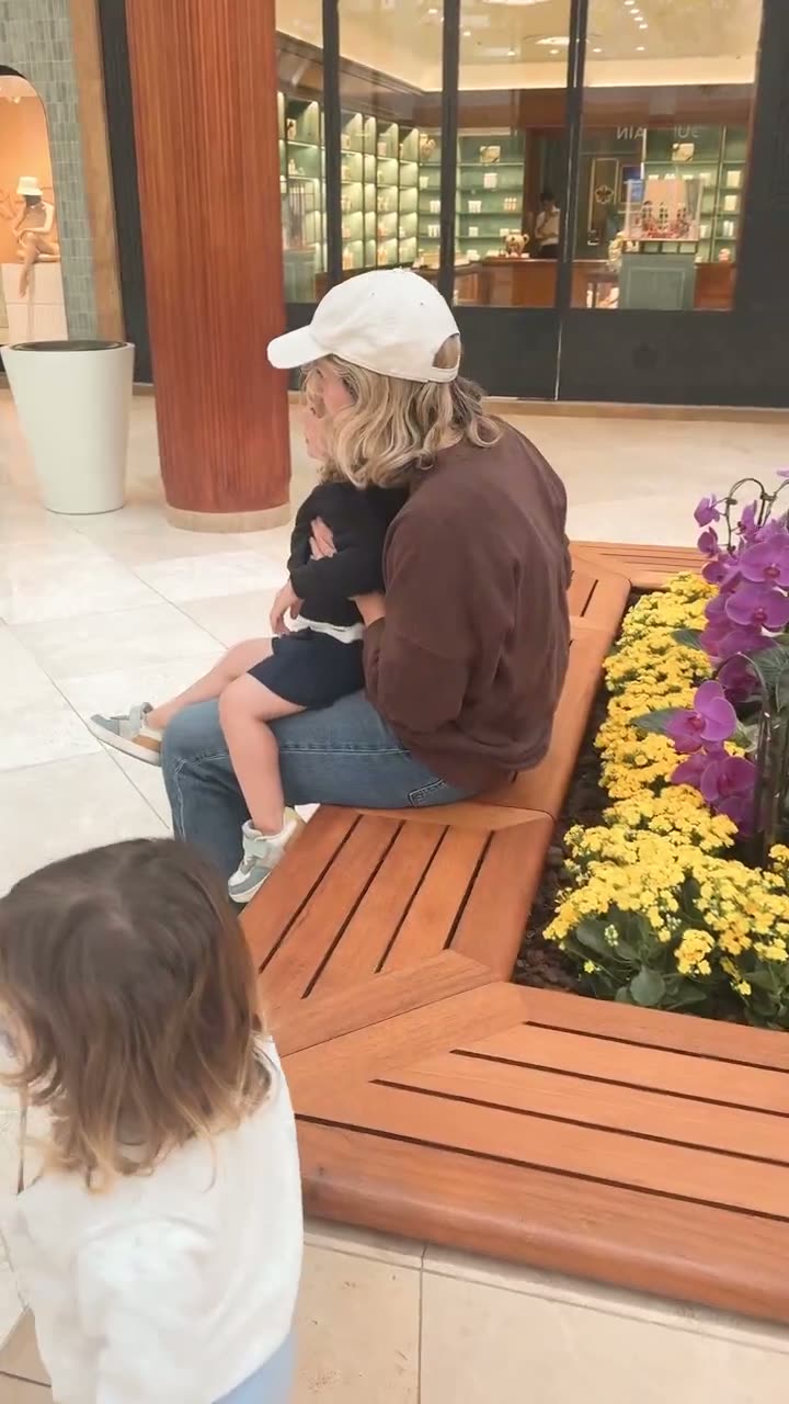 A short clip at South Coast Plaza of a toddler held on a bench beside the planters while the second twin waits nearby, just after a replacement balloon was bought