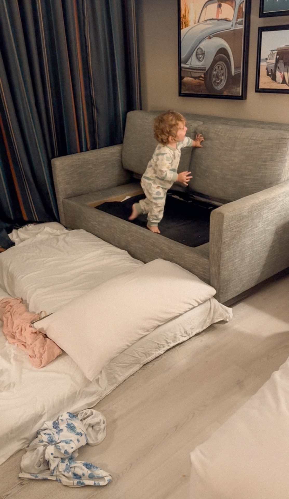 A small child standing inside an opened hotel sofa bed, with bedding and pillows spread across the floor