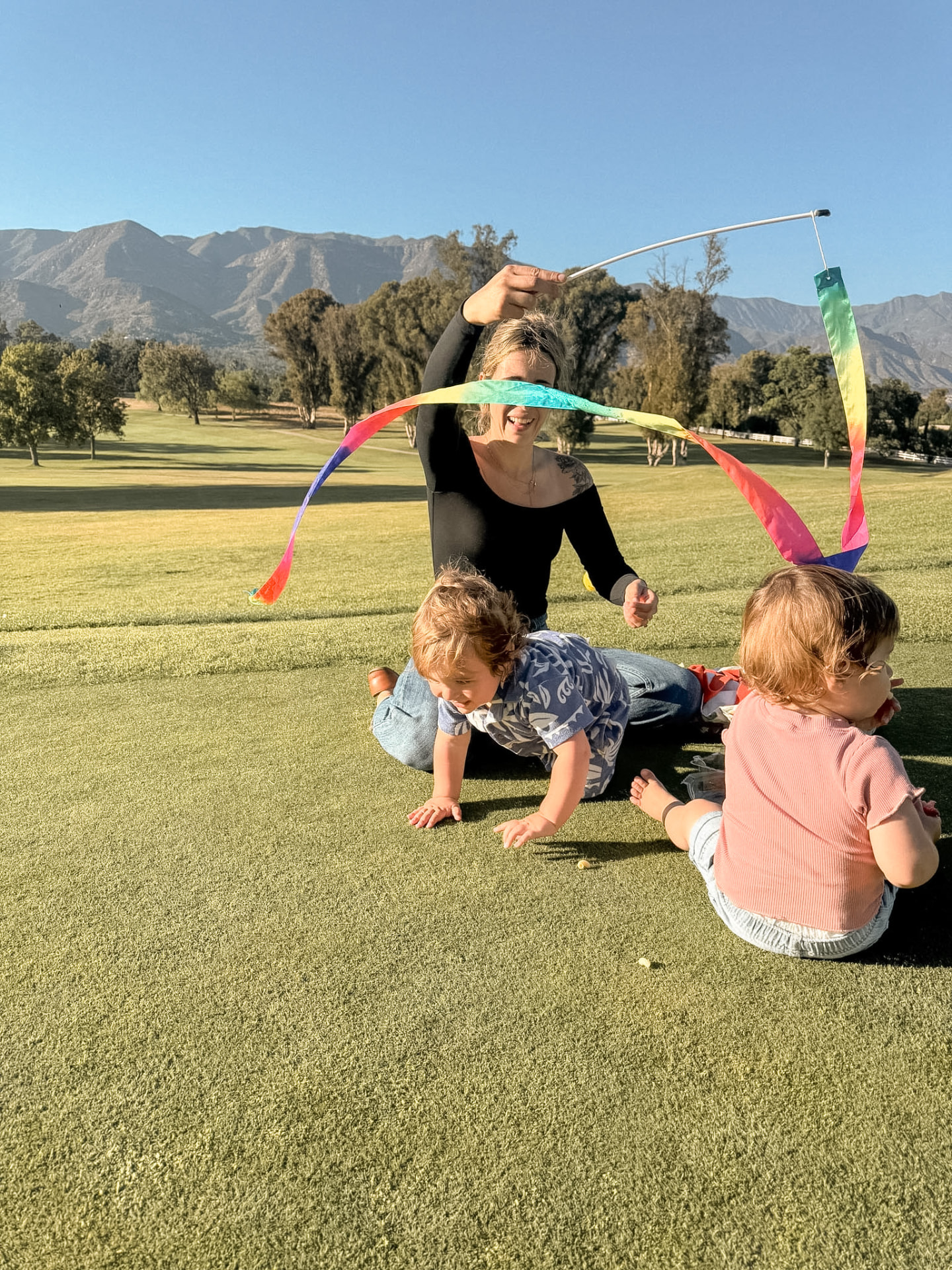 A mother waving a long ribbon wand for two toddlers on a wide lawn with the Ojai mountains behind