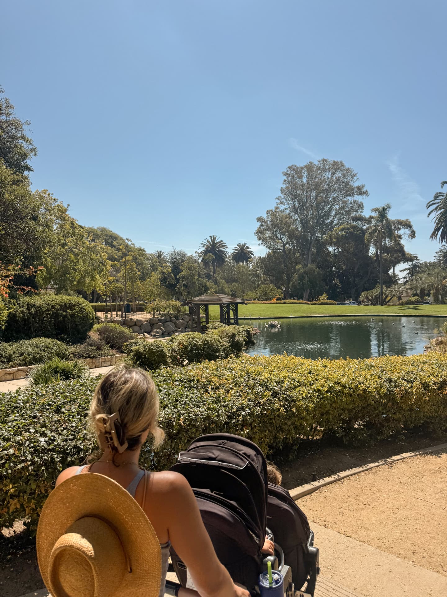A parent pushing a double stroller along a shaded path beside the duck pond at Alice Keck Park in Santa Barbara