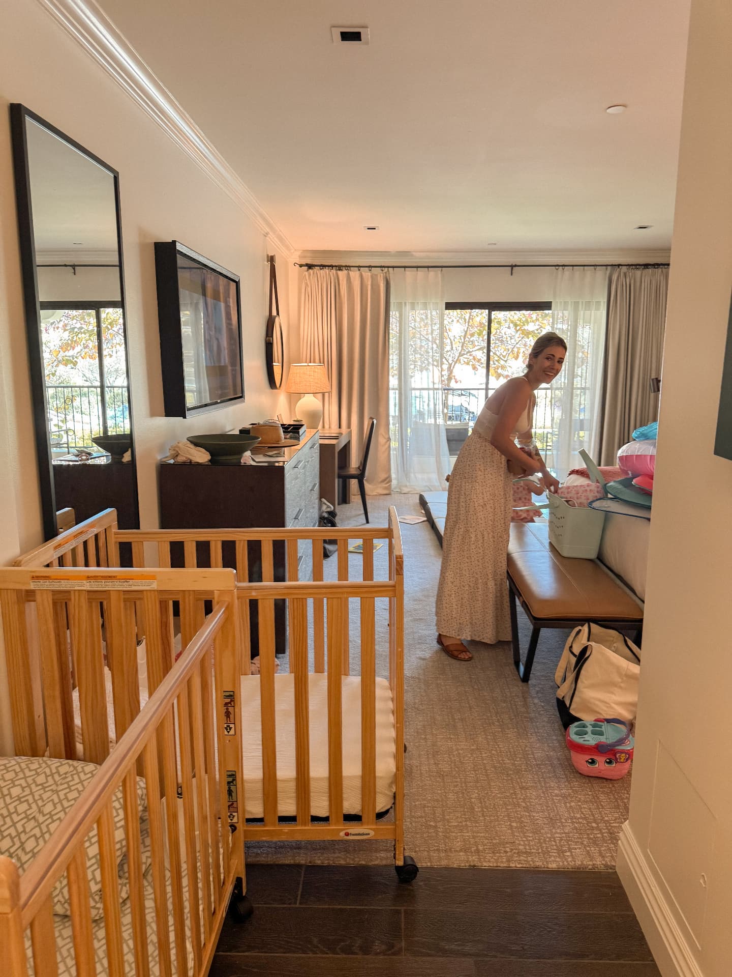 A hotel room in use, with two wooden travel cots, bags and children's things spread across the floor while a parent unpacks by the window