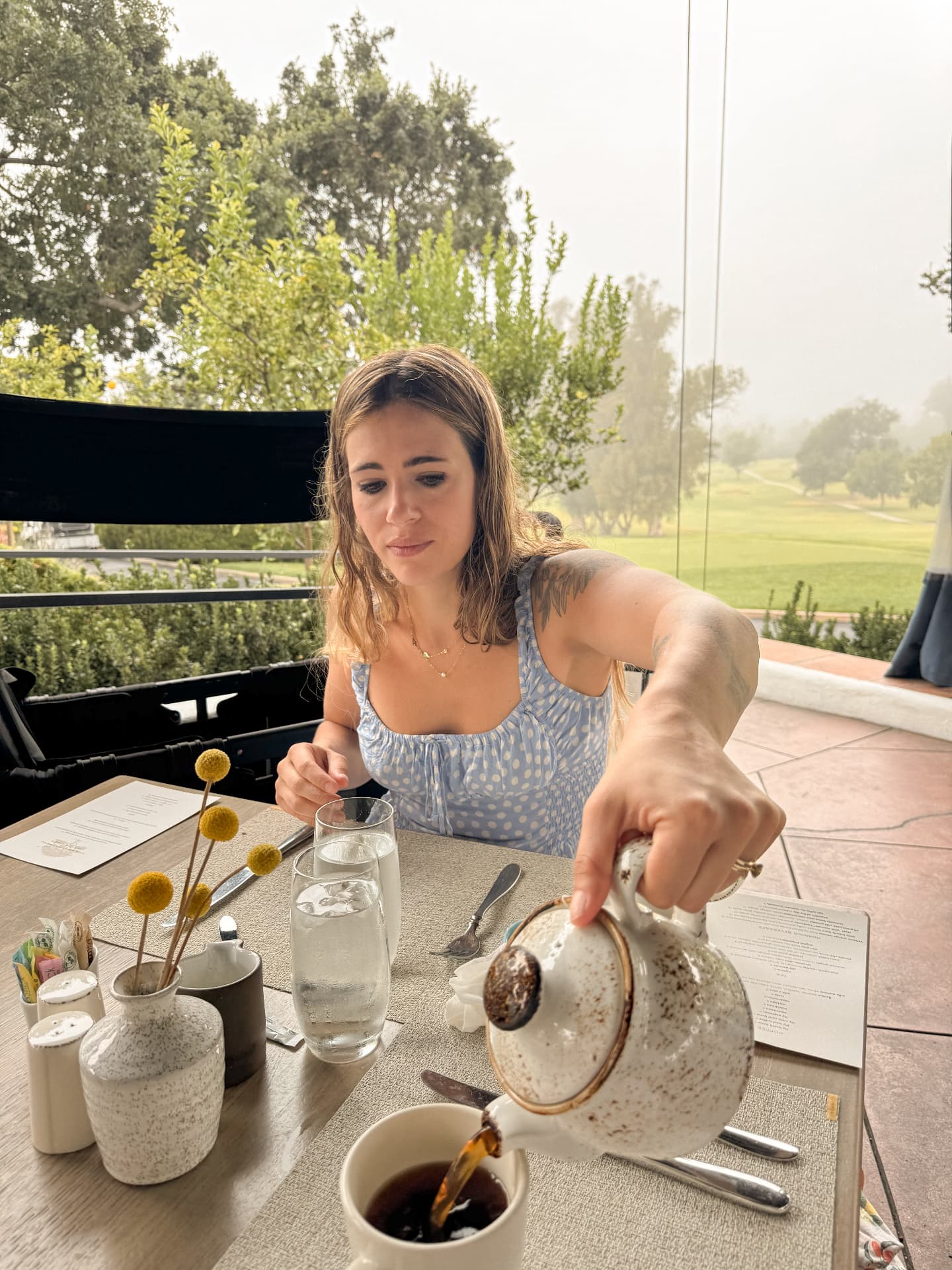 Linda pouring tea from a stoneware pot at a breakfast table at The Oak, misty golf course beyond the windows
