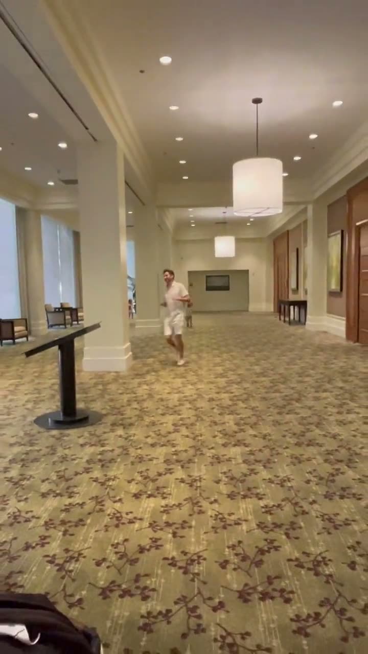 A short clip of two toddlers running the length of a large, empty hotel banquet room with patterned carpet and drum-shade lighting