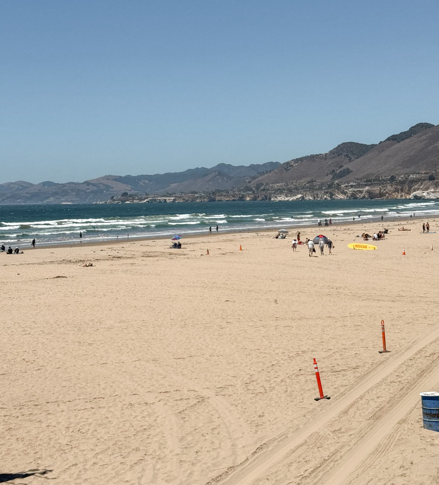 The wide flat sand of Pismo Beach on a clear summer day, with surf, a few families and the coastal hills in the distance