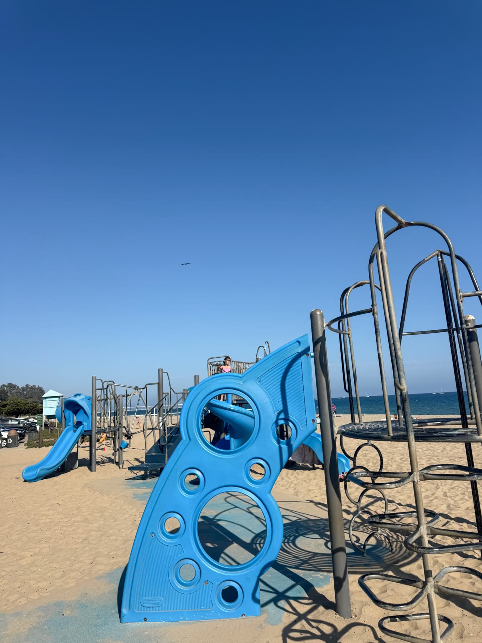 A blue and grey playground structure standing in the sand beside the beach in Santa Barbara, with the ocean behind it
