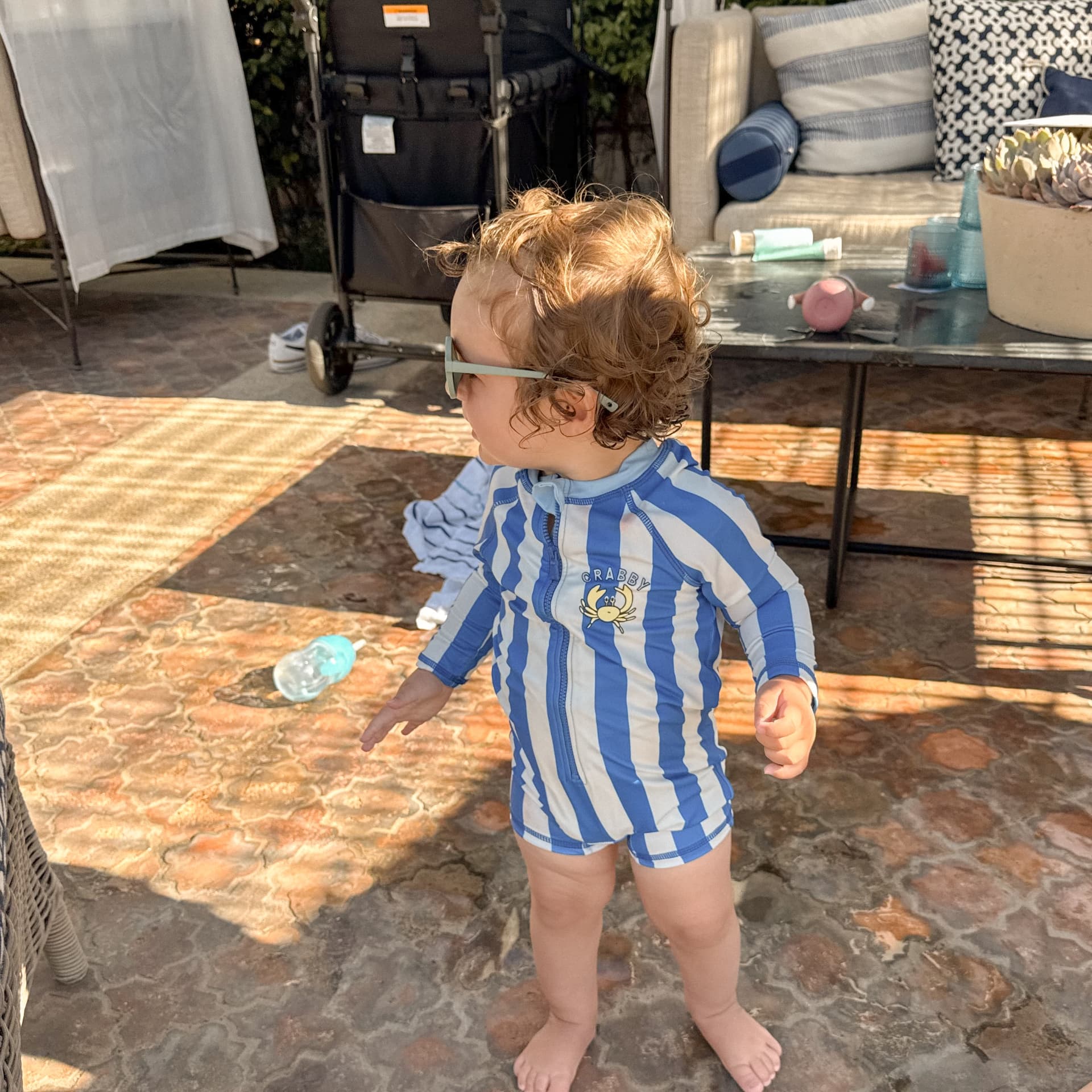 A toddler in a blue striped swimsuit and sunglasses standing on the tiled patio beside the pool