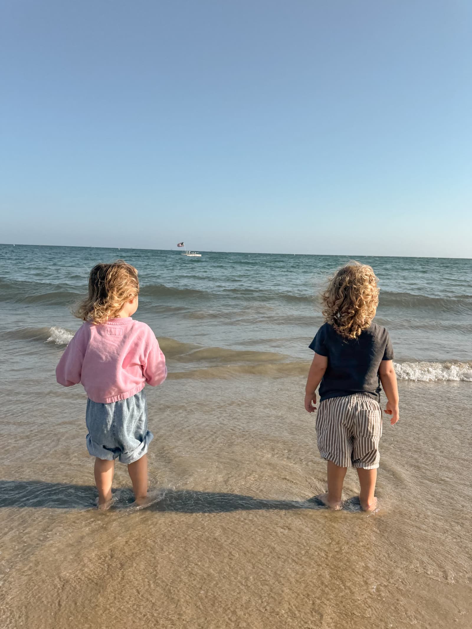 Two toddlers standing ankle-deep in the ocean with their backs to the camera, looking out at the water on a clear afternoon