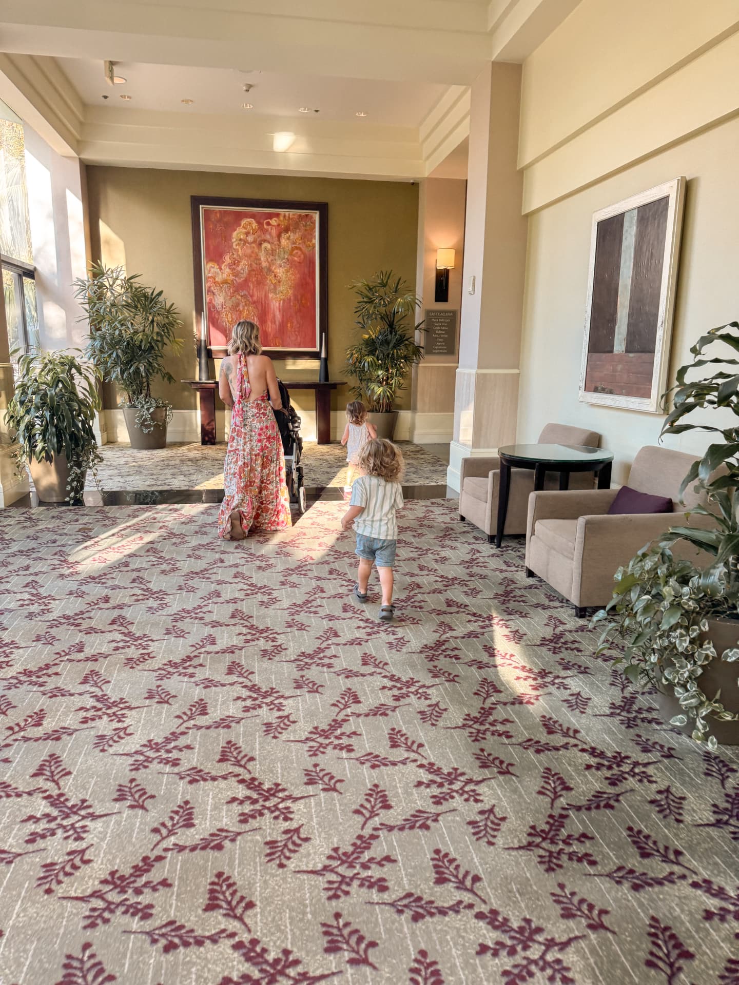 Linda walking through a hotel lobby pushing a stroller while two-year-old twins August and Amelia walk behind her