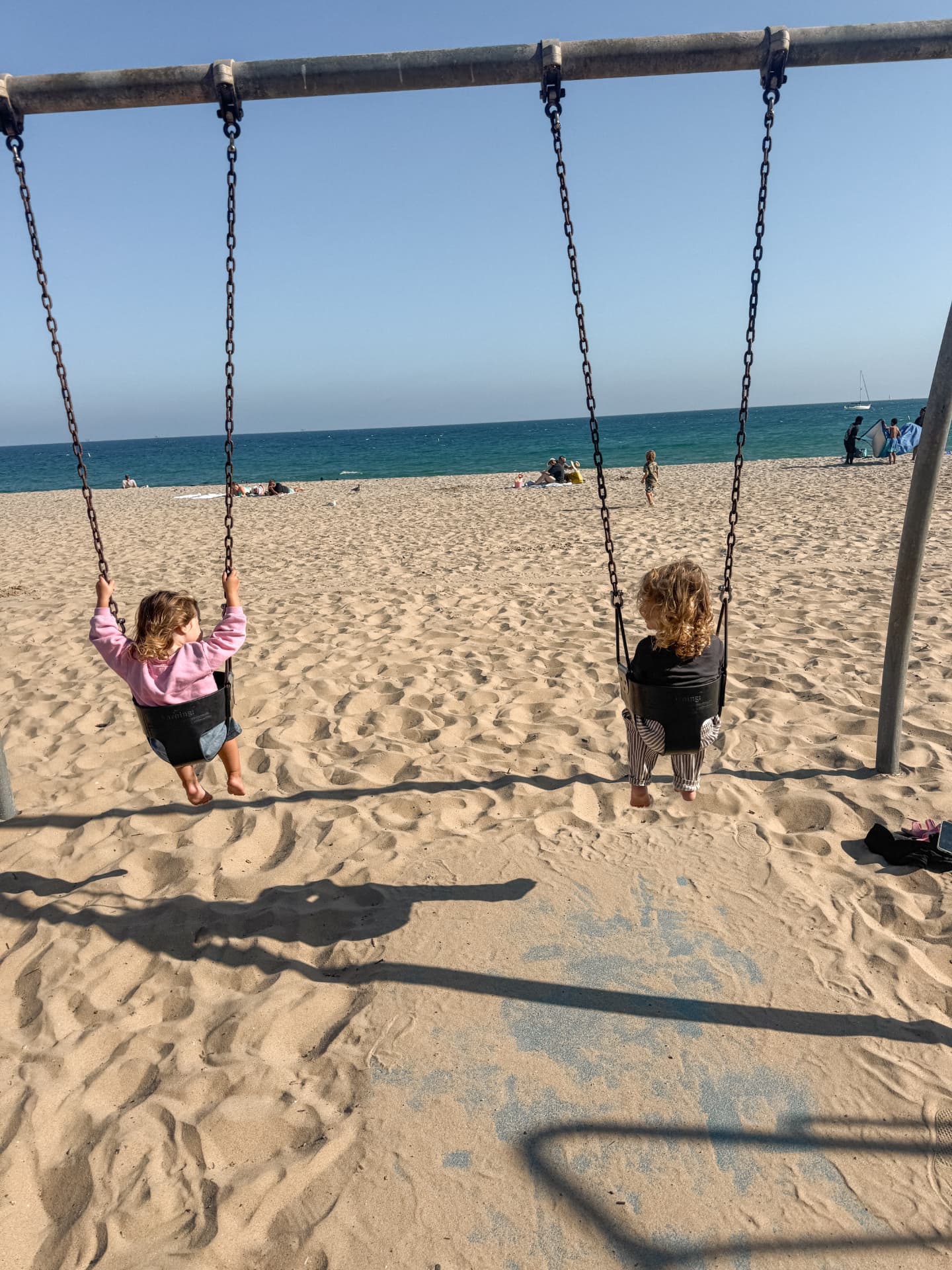 Two toddlers swinging side by side on a beach swing set in the sand, facing the ocean in Santa Barbara
