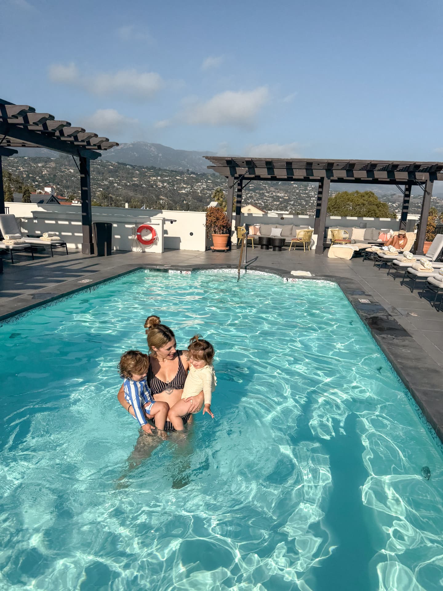 Linda standing in the rooftop pool at the Kimpton Canary Hotel holding both 16-month-old twins, with pergolas and the Santa Barbara foothills behind them
