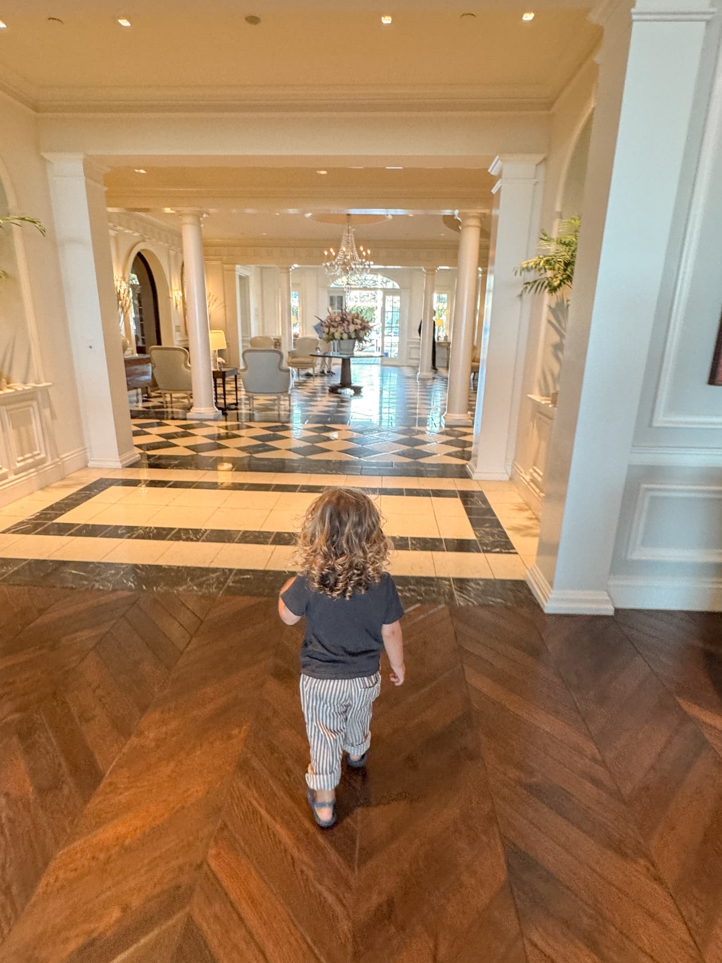 A small child walking alone across a chequered marble floor toward a bright hotel lobby