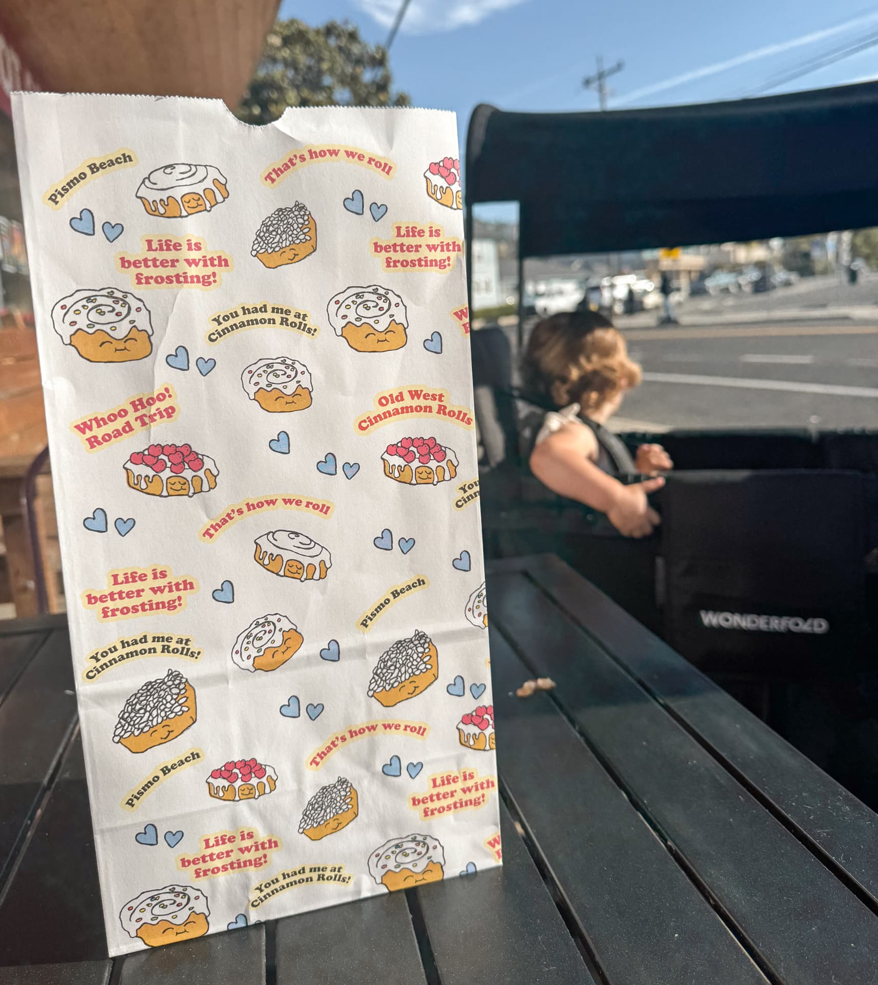 An Old West Cinnamon Rolls paper bag on an outdoor table with one of our twins sitting in the stroller behind it